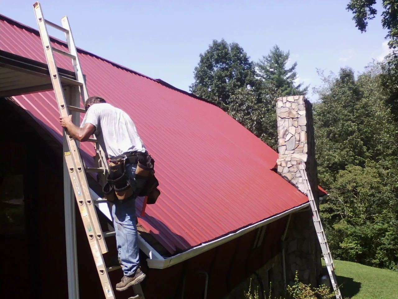 Roofer on a ladder beside a red metal roof and stone chimney — Soffit & Fascia Repair by Roofing Brothers in Mannington